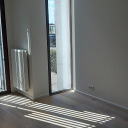 Marcin Szarek - Jasne wnętrze po remoncie: świeżo pomalowane ściany, drewniana podłoga, grzejnik i okno z widokiem na zieleń. Promienie słońca tworzą geometryczne wzory na podłodze.