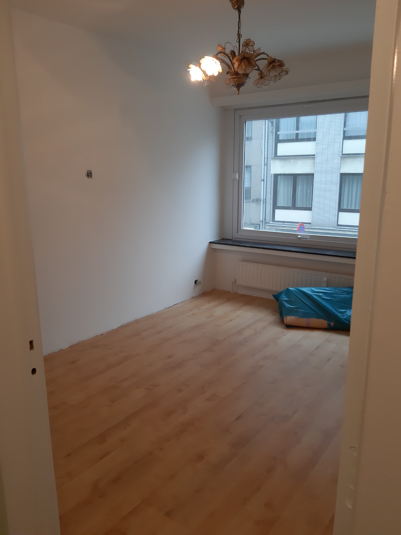 Pusty pokój po remoncie z drewnianą podłogą, białymi ścianami i oknem z widokiem na budynek. Pod oknem grzejnik i zabezpieczone folią meble. W tle ozdobny żyrandol.