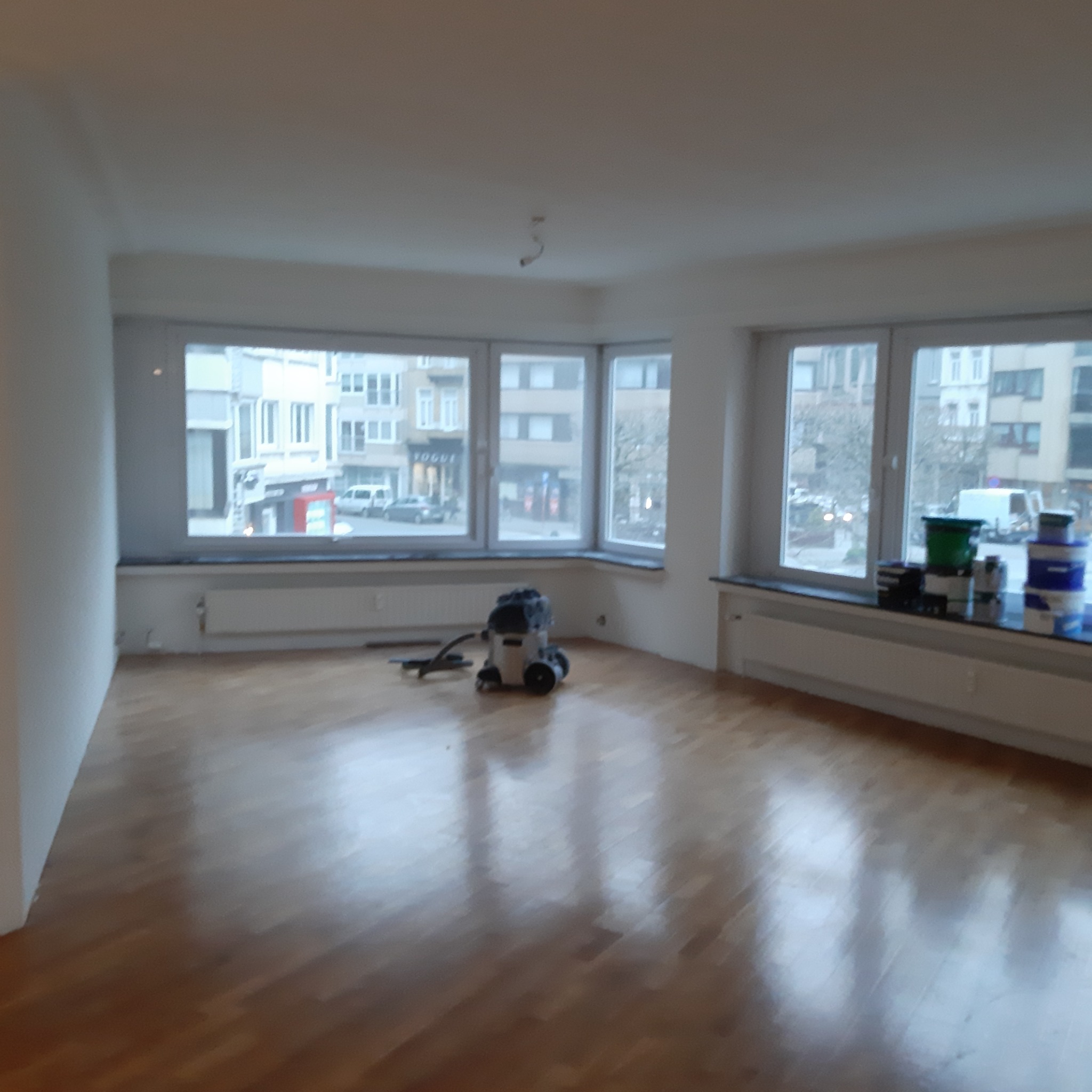 Pusty pokój po remoncie z odkurzaczem i farbami przy oknie. Parkiet odnowiony, ściany przygotowane do malowania. Widok na ulicę z budynkami w tle.