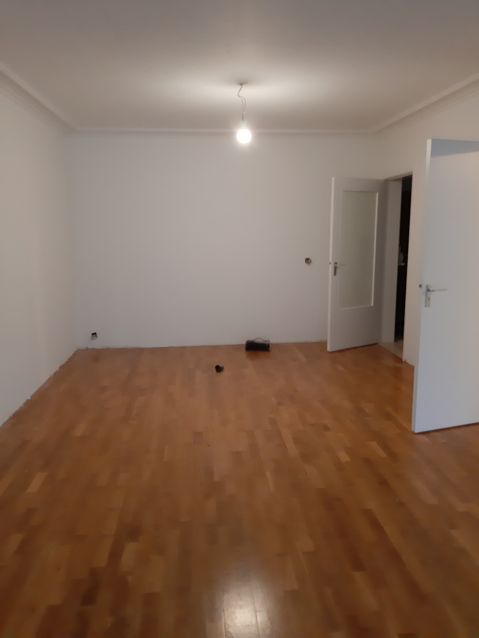 Pusty pokój z drewnianą podłogą i białymi ścianami. Widoczne otwarte drzwi i listwy przypodłogowe. Minimalistyczne wnętrze przed wykończeniem.