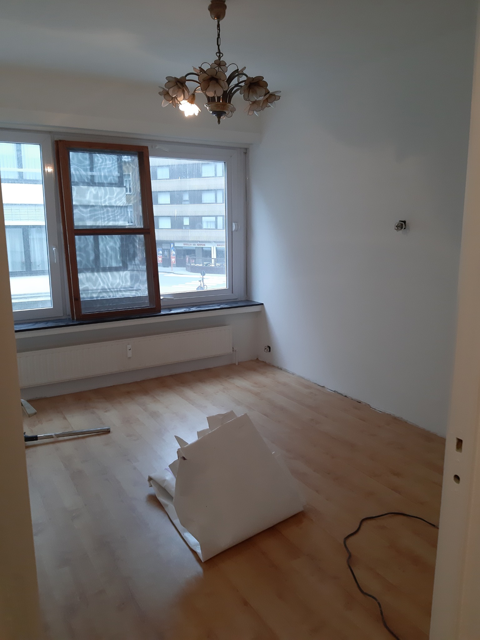 Pokój przed remontem: gołe ściany, drewniana podłoga, okno z brązową ramą, zwisający żyrandol. Widoczne narzędzia i resztki materiałów budowlanych.