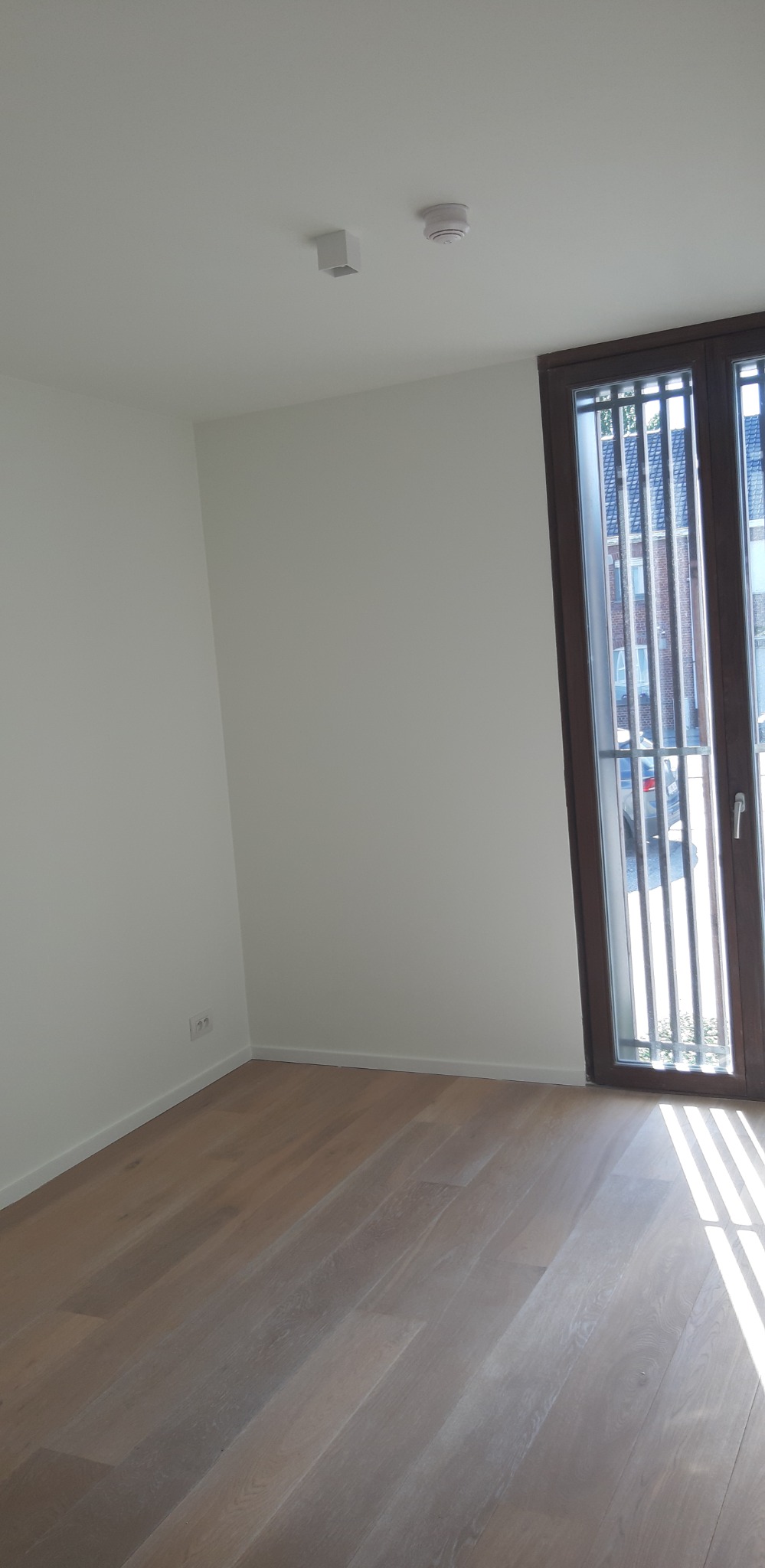 Jasne, puste wnętrze po remoncie z drewnianą podłogą, białymi ścianami i oknem z ciemną ramą. Widoczne detale wykończeniowe, takie jak listwy przypodłogowe.