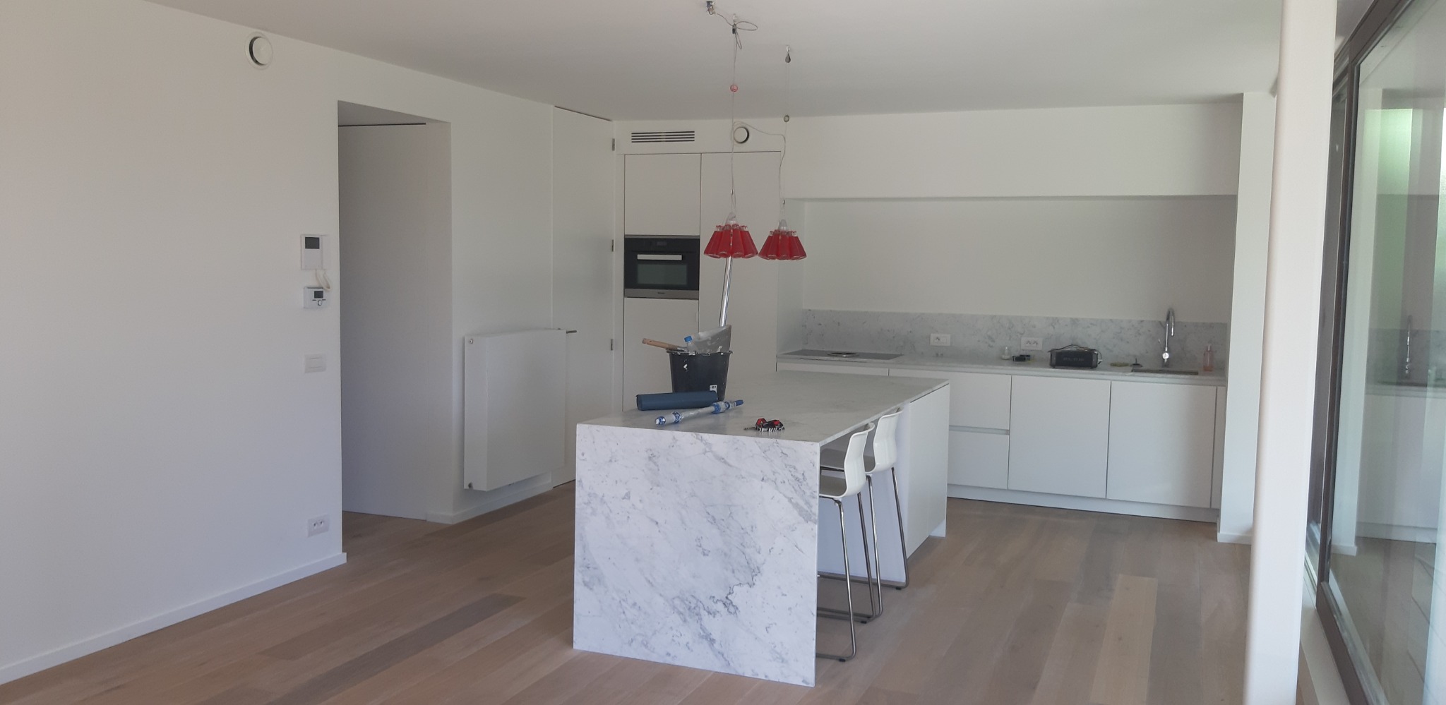 Jasne wnętrze kuchni z wyspą z marmurowym blatem, widoczne narzędzia malarskie sugerują etap wykończeniowy. Minimalistyczny design, białe ściany i drewniana podłoga.