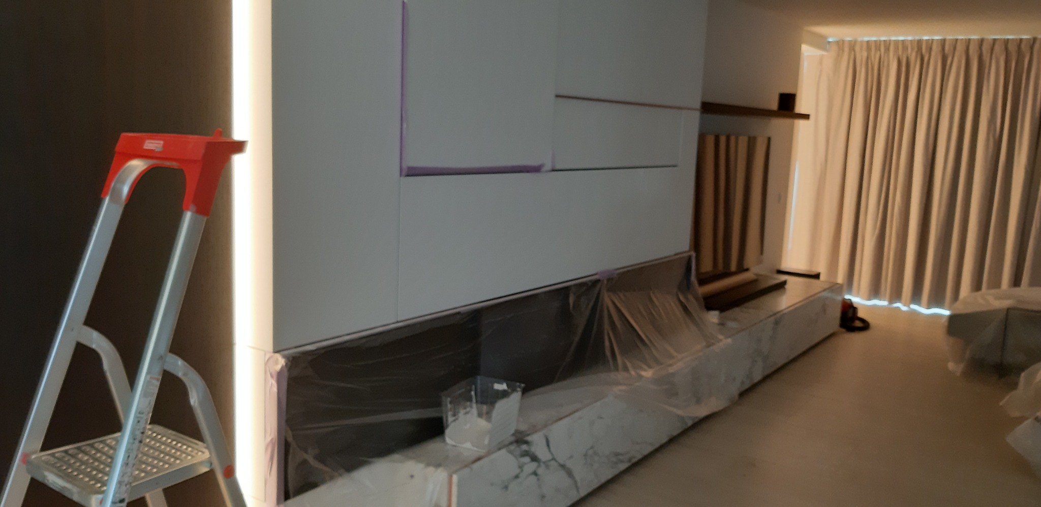 Biała ściana z podświetleniem LED w trakcie malowania, zabezpieczona folią. Widoczna drabina malarska i wiaderko z farbą. Wykończenie wnętrz w nowoczesnym stylu.