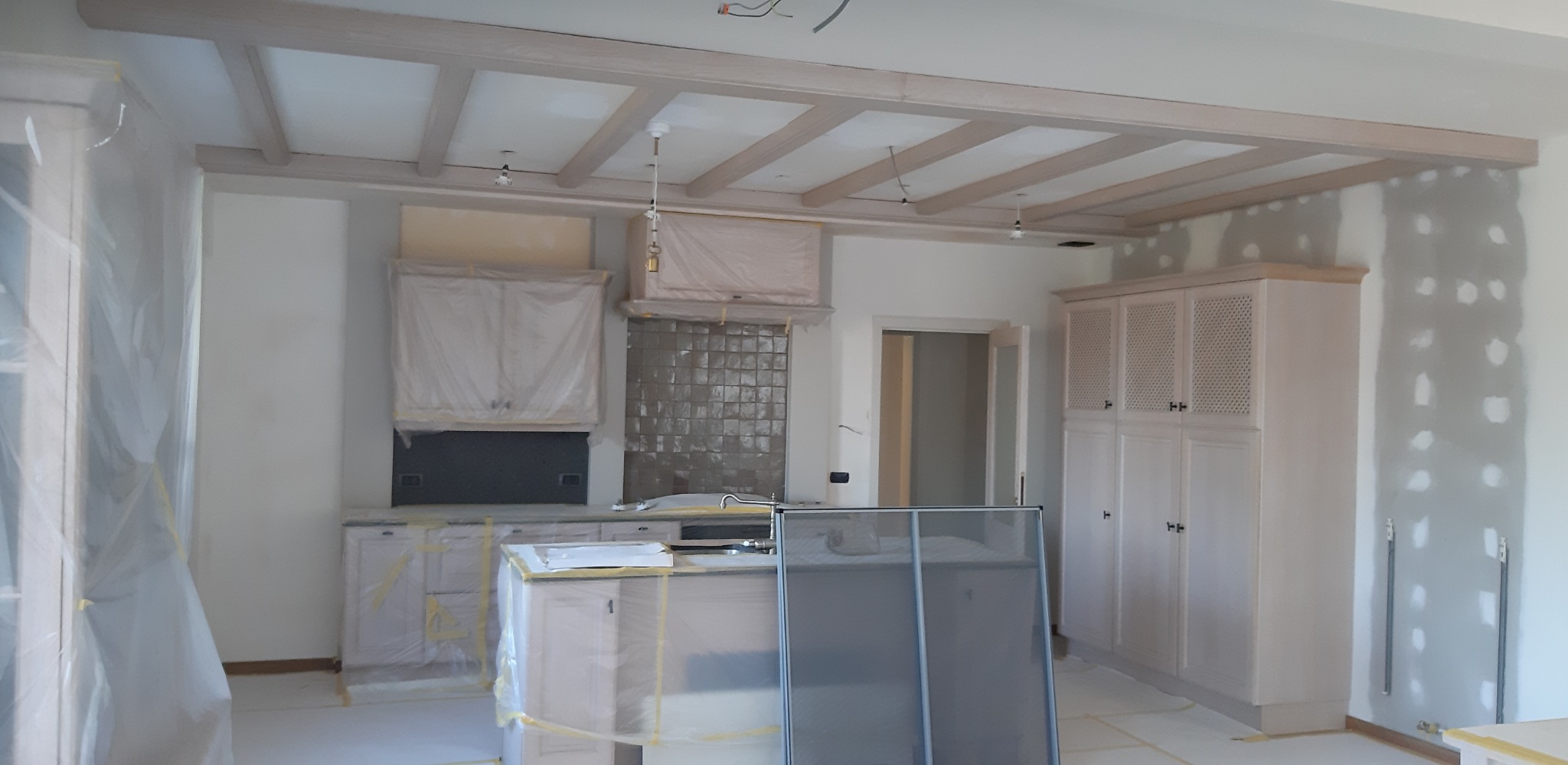 Kuchnia w trakcie prac wykończeniowych: zabezpieczone meble folią, sufit z drewnianymi belkami, ściany częściowo wyszpachlowane, widoczne instalacje elektryczne.