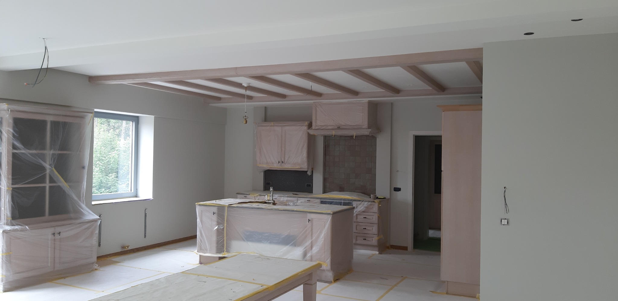 Wnętrze kuchni w trakcie prac wykończeniowych, meble zabezpieczone folią, widoczne drewniane elementy dekoracyjne pod sufitem i świeżo pomalowane ściany w jasnym odcieniu.