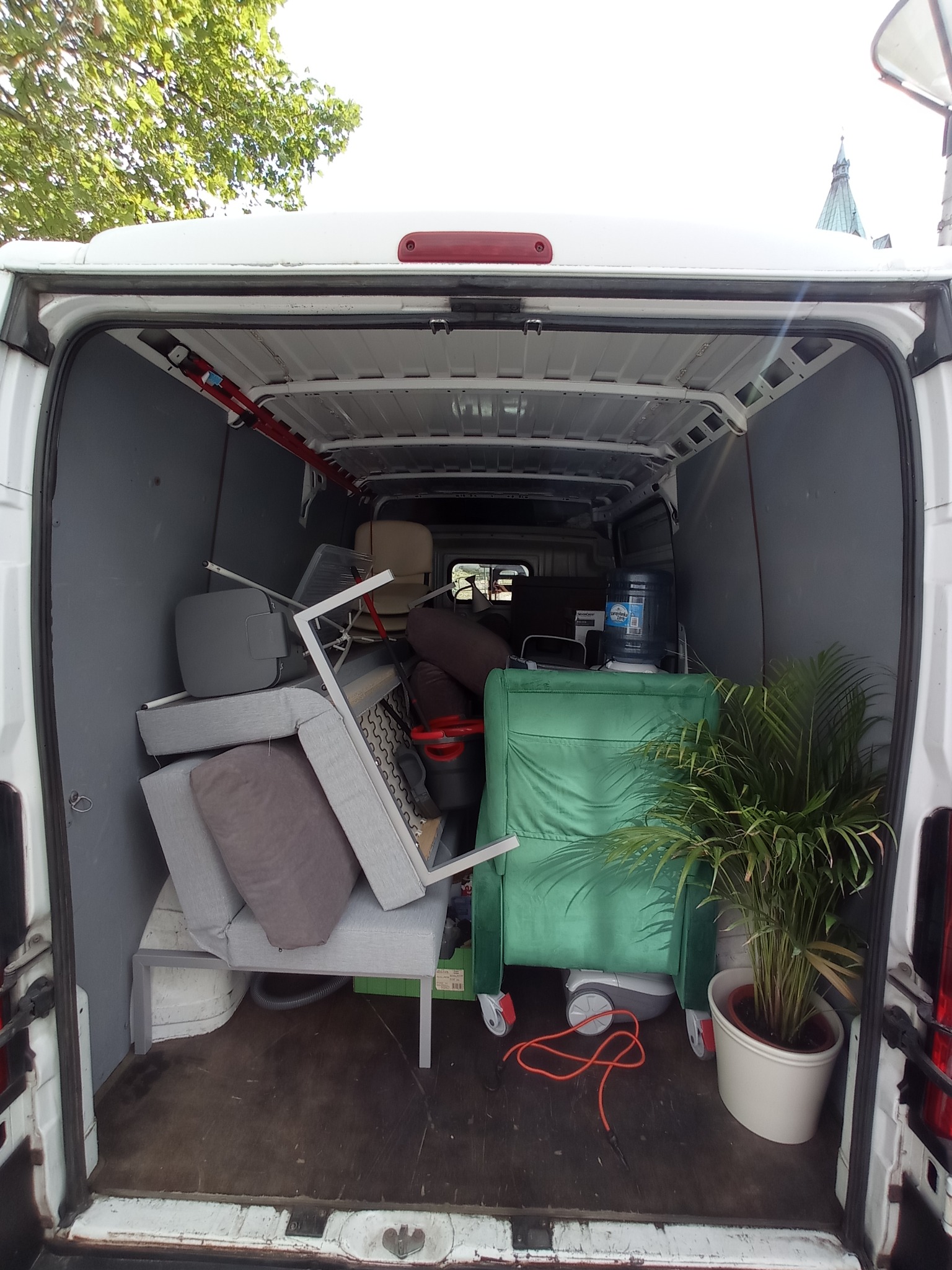 Wnętrze białego busa załadowane meblami, krzesłami, rośliną doniczkową i dystrybutorem wody. Widok z tyłu pojazdu, podczas załadunku. Część przedmiotów nachodzi na siebie.