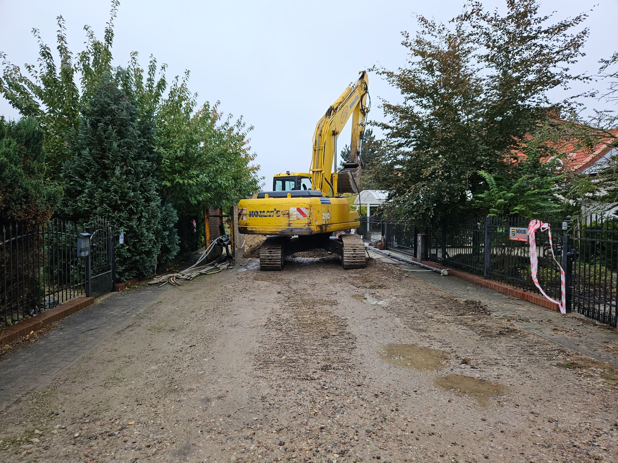 Żółta koparka Komatsu 210 na drodze wjazdowej, otoczona zielenią i ogrodzeniem, przygotowuje teren pod budowę. Widok z tyłu maszyny.