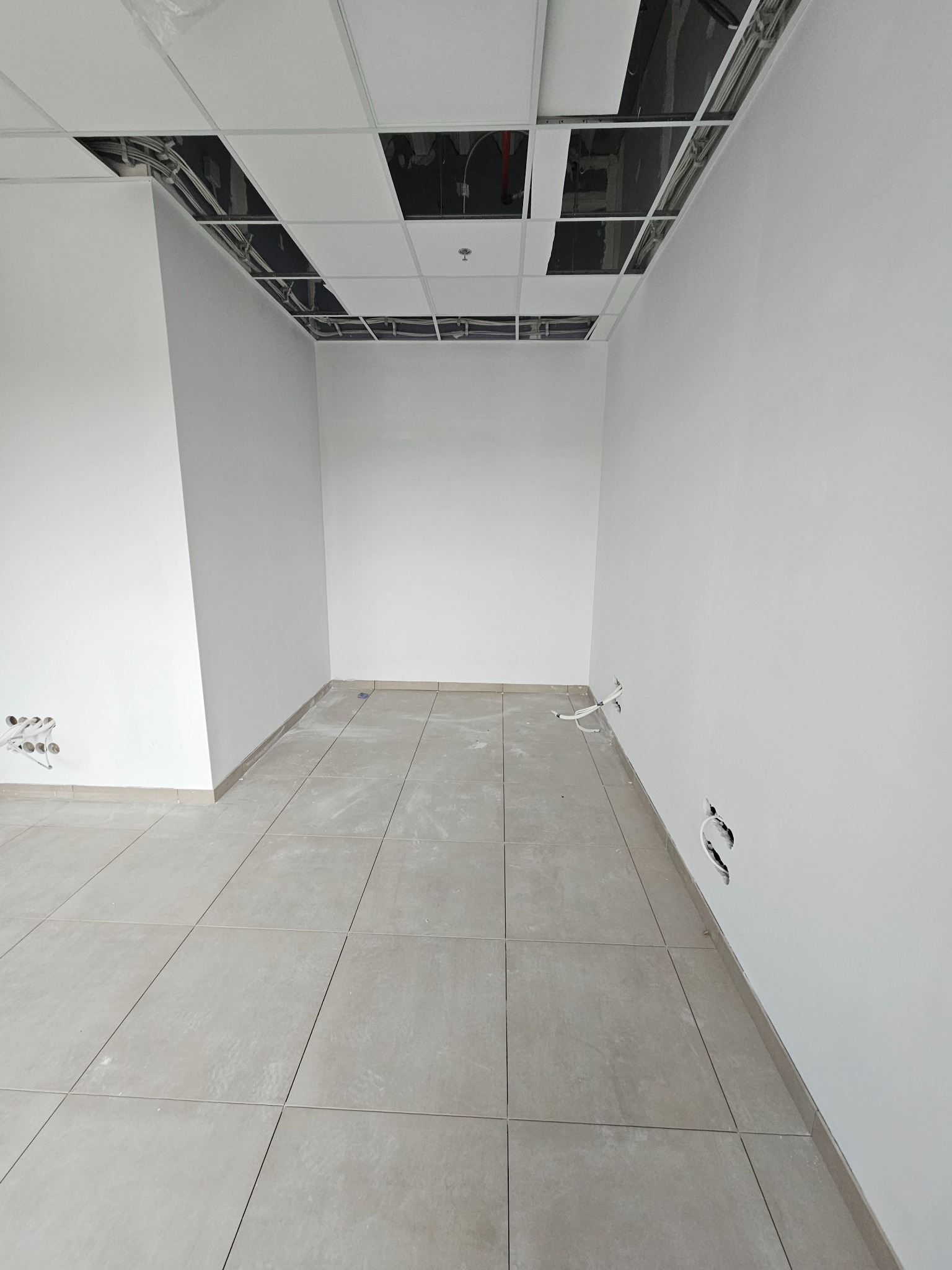 Wnętrze w trakcie wykończenia: szare płytki podłogowe, białe ściany, sufit podwieszany z widoczną instalacją. Widoczne gniazdka elektryczne i otwory na kable.