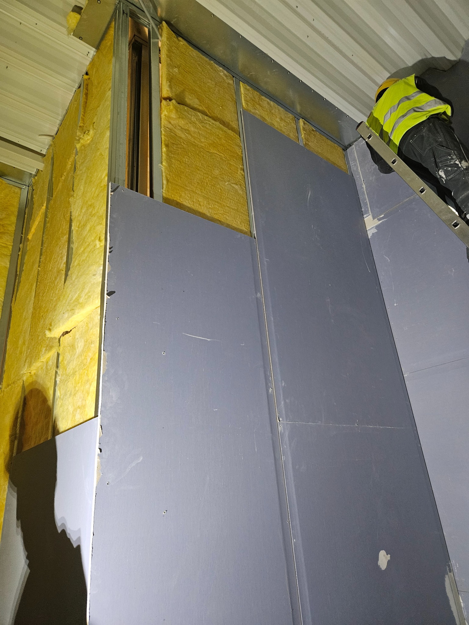 Montaż płyt gipsowo-kartonowych na ścianie z izolacją z wełny mineralnej. Widoczny fragment drabiny i pracownik w kamizelce odblaskowej. Ujęcie z dołu.