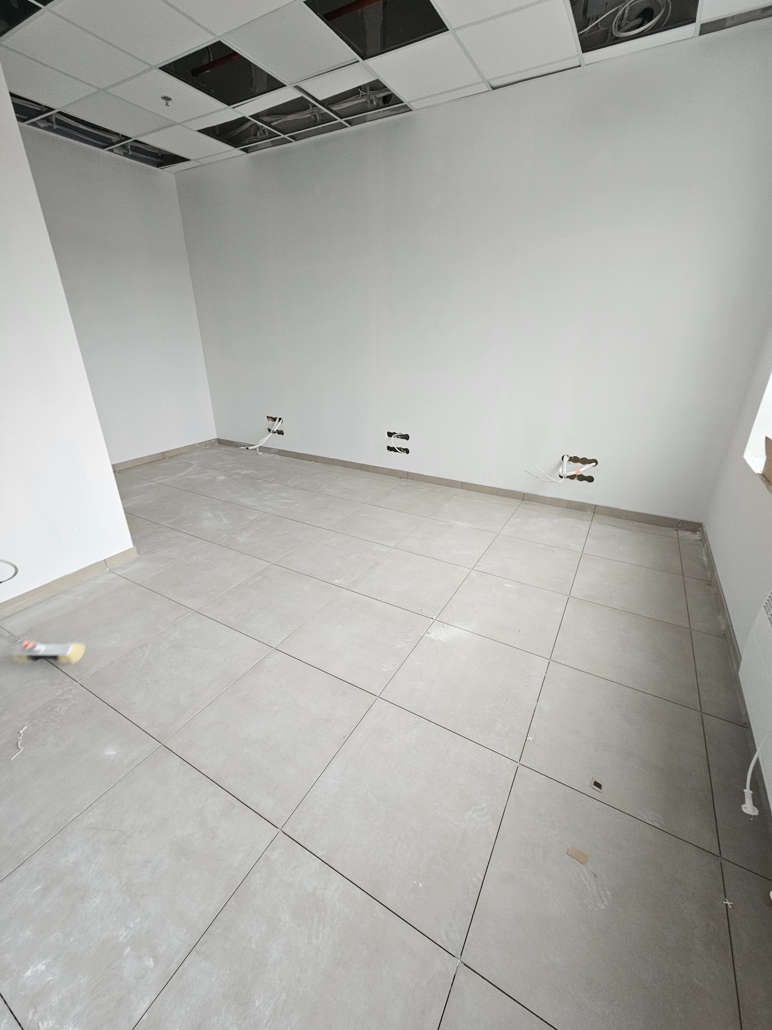 Wykończone płytki podłogowe w jasnym odcieniu szarości w pustym pomieszczeniu z widocznymi instalacjami elektrycznymi i fragmentem sufitu podwieszanego.