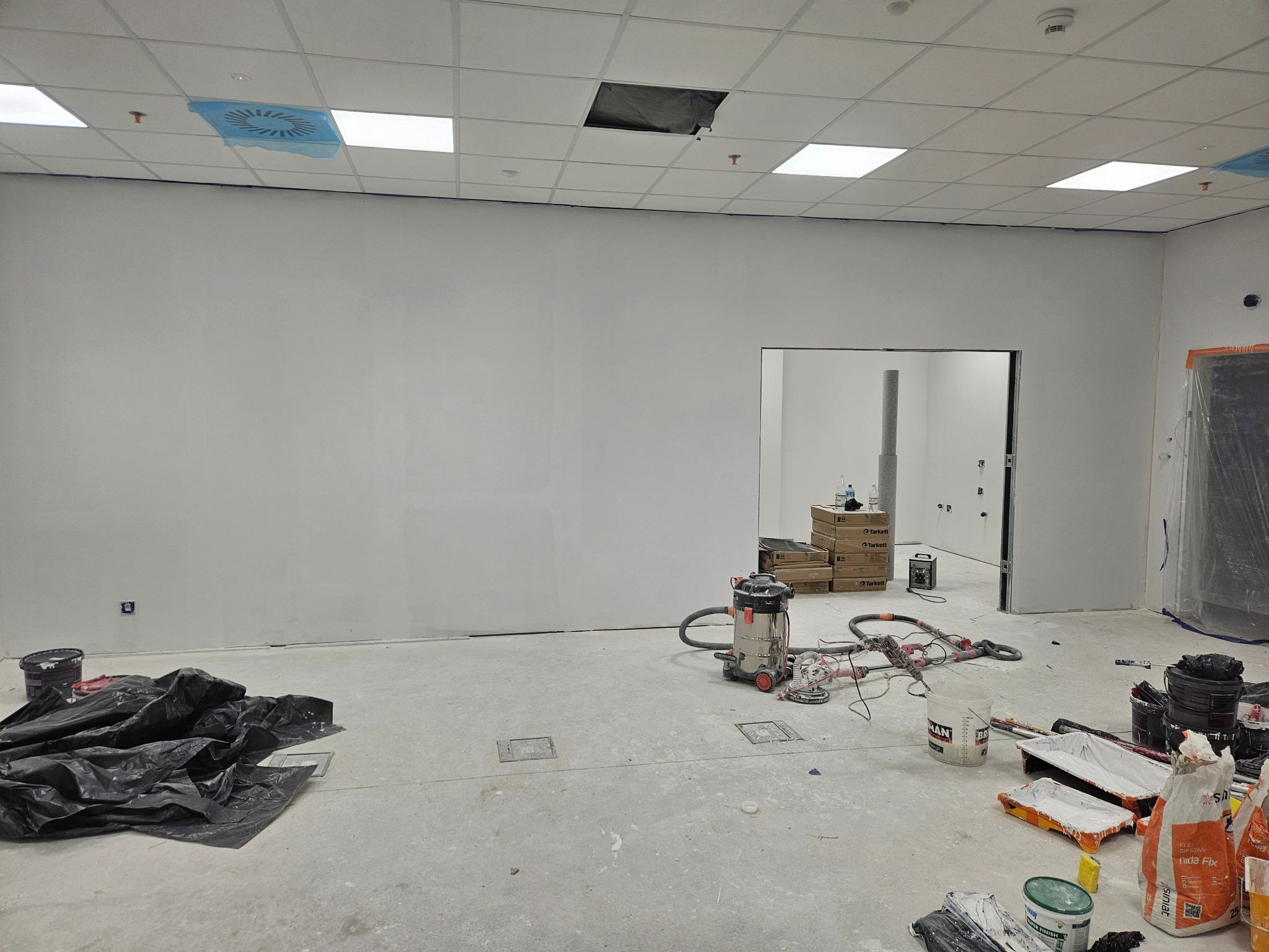 Przygotowania do malowania ścian w pomieszczeniu: zakryte elementy, narzędzia malarskie, szpachla, odkurzacz przemysłowy. Widoczne ślady szpachlowania na ścianach.