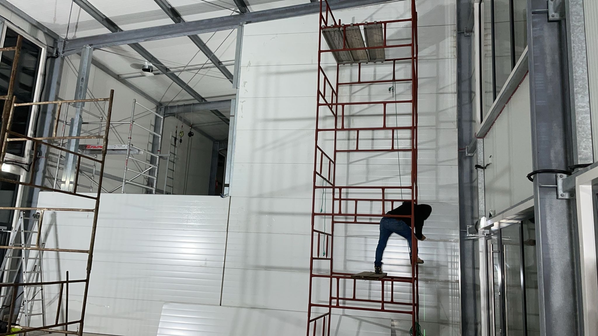 Montaż paneli ściennych w hali przemysłowej. Pracownik na rusztowaniu mocuje białe panele. Widoczne metalowe konstrukcje i oświetlenie sufitowe.