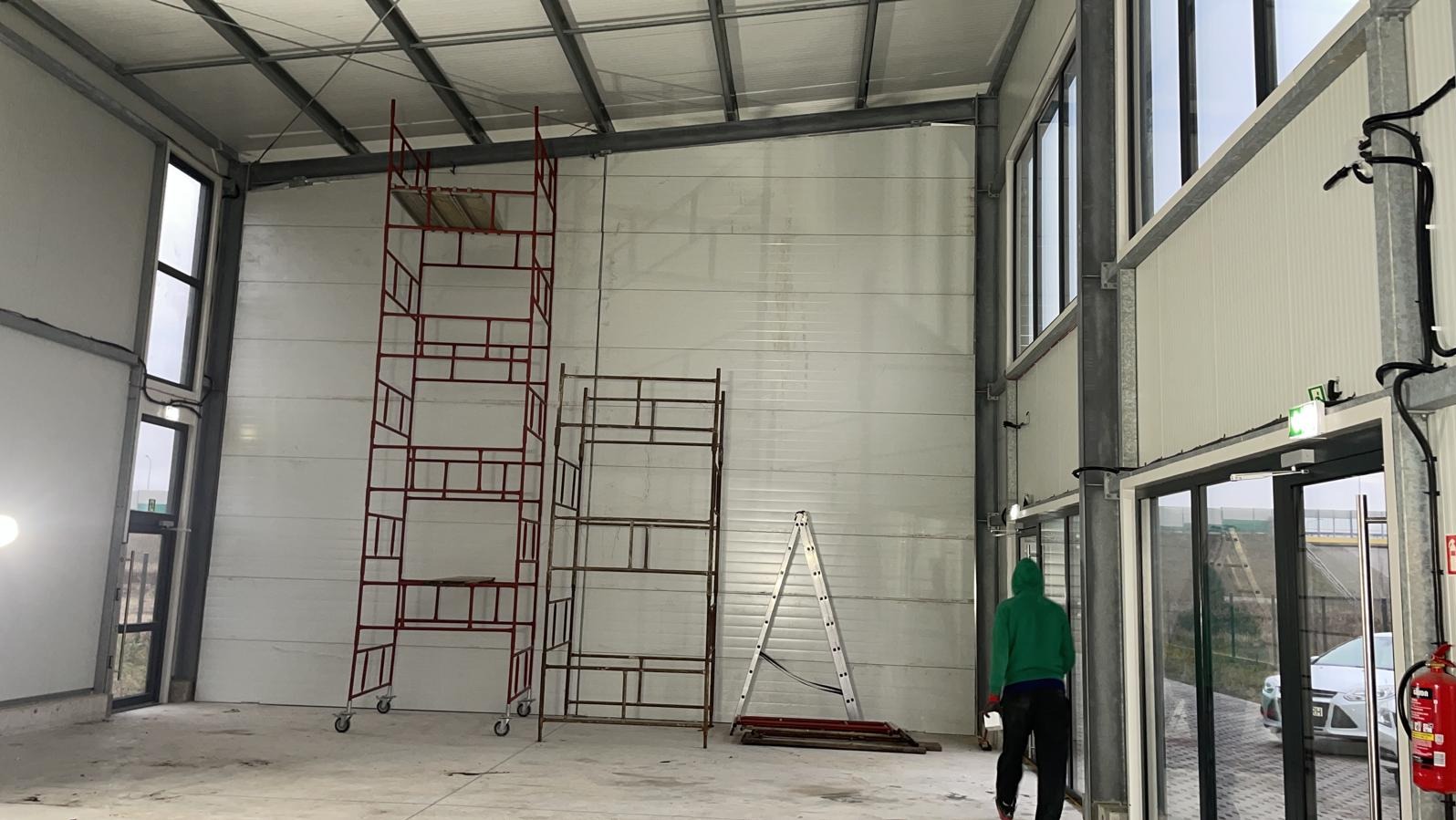 Wnętrze hali produkcyjnej w trakcie wykańczania: białe ściany z blachy trapezowej, stalowa konstrukcja, rusztowania, drabina i osoba w zielonej bluzie przy drzwiach.