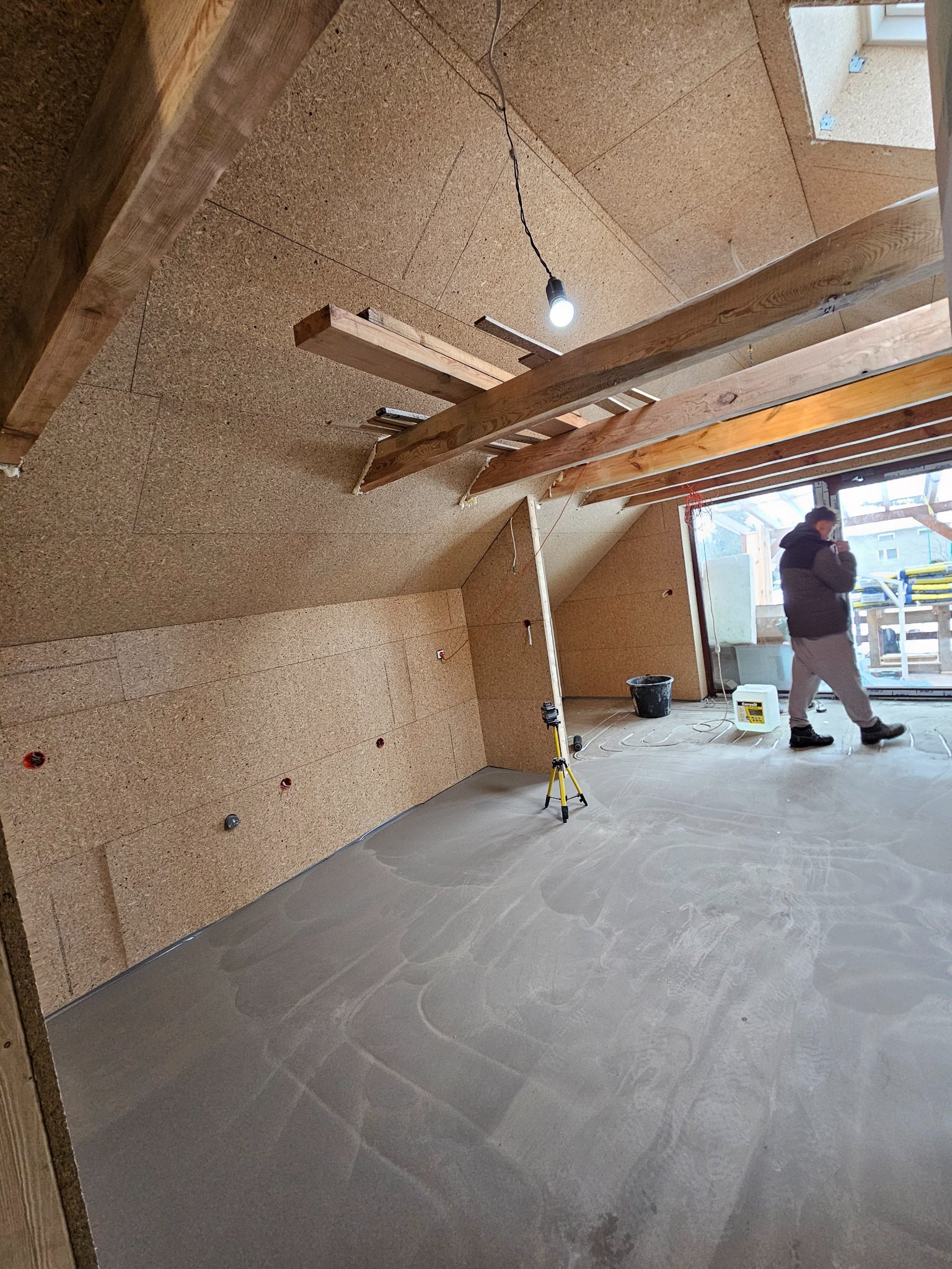 Poddasze w trakcie prac wykończeniowych: ściany obłożone płytami OSB, widoczne drewniane belki stropowe, wylewka na podłodze, osoba w tle. Oświetlenie robocze.