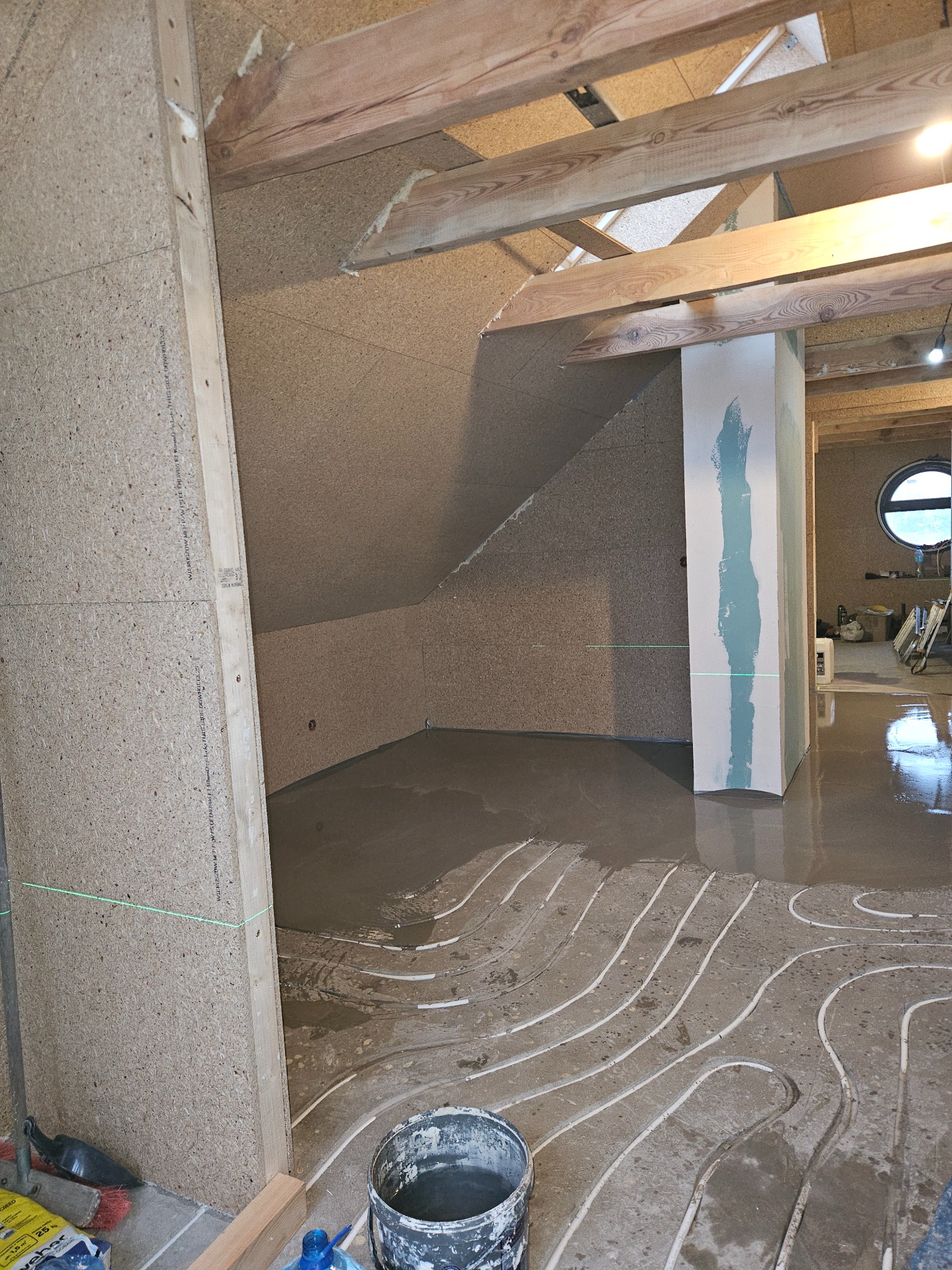 Podłogówka w szarej wylewce, widoczne rury grzewcze. Ściany wyłożone płytami OSB, sufit z drewnianych belek. W tle okrągłe okno i narzędzia.