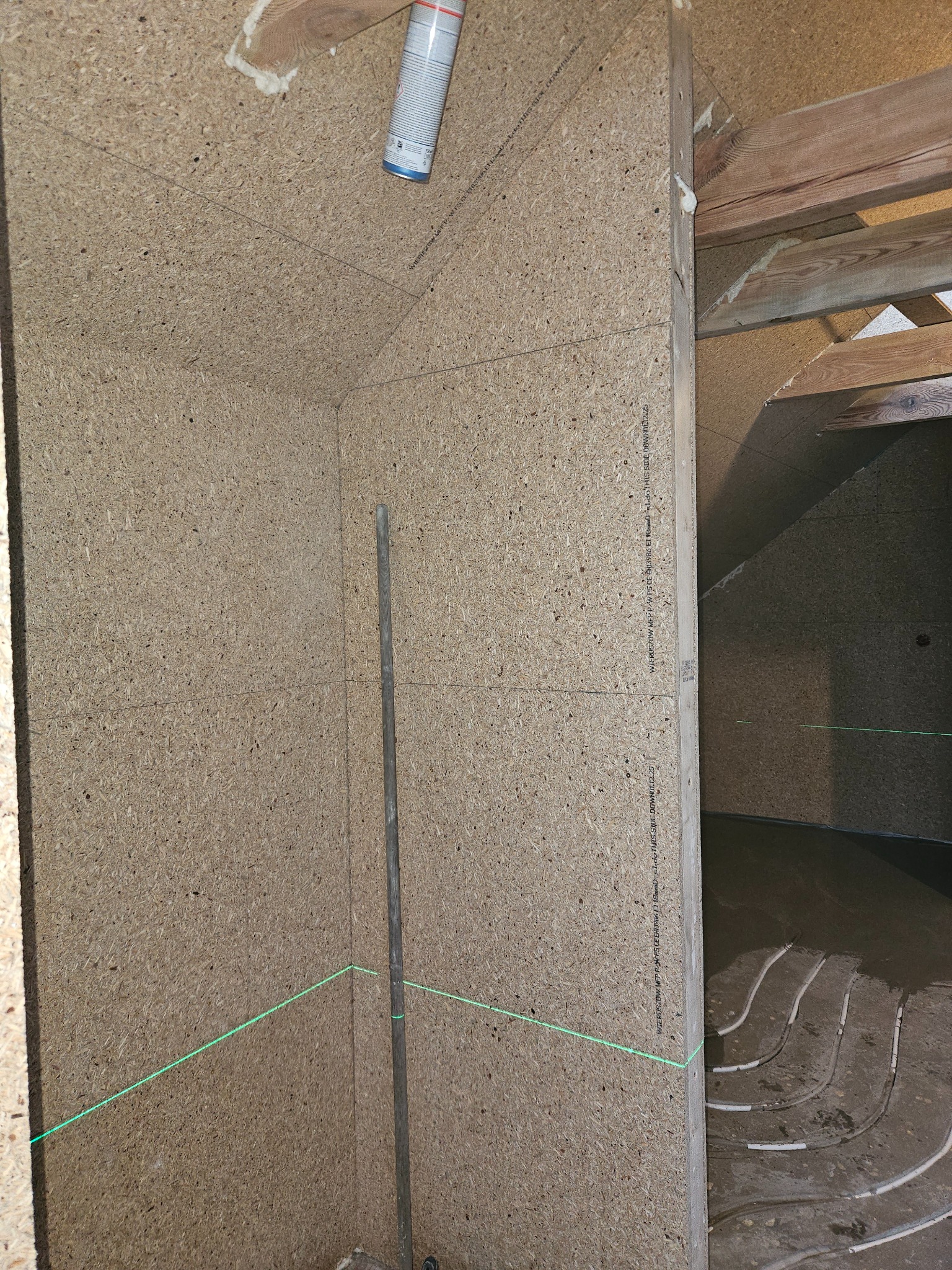 Montaż płyty drewnopochodnej na poddaszu z widoczną instalacją ogrzewania podłogowego. Poziomica laserowa wyznacza linię. Puszka pianki montażowej w rogu.