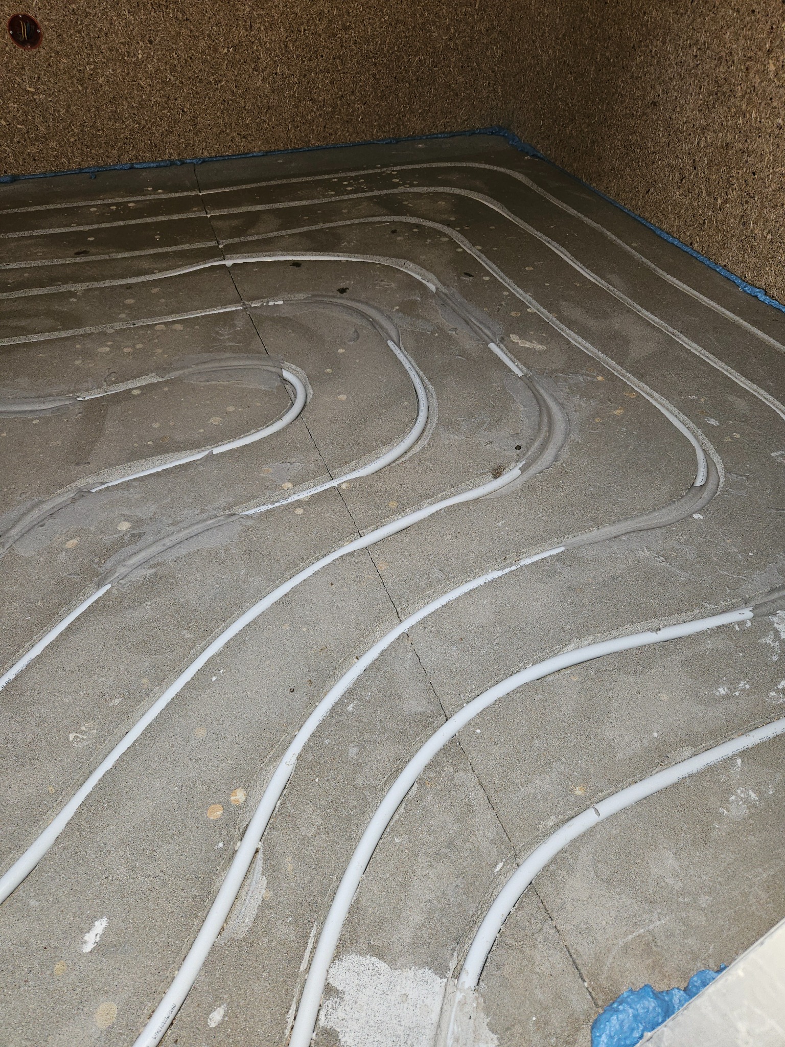 Instalacja ogrzewania podłogowego w trakcie prac w Warszawie. Białe rurki ułożone w betonowej wylewce, widoczne fragmenty izolacji przy ścianach.