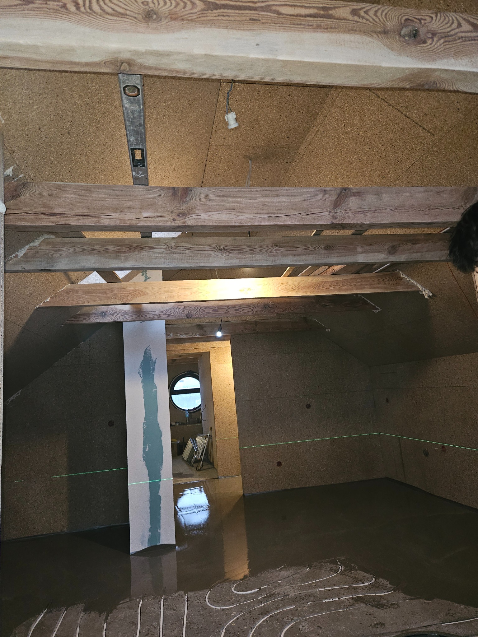 Poddasze w trakcie prac wykończeniowych: widoczne belki stropowe, płyty OSB na ścianach, wylewka na ogrzewaniu podłogowym, poziomica laserowa i okrągłe okno w tle.