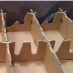 Wnętrze kartonowego opakowania z przegródkami, widok z góry. Brązowy karton, szare krawędzie, otwory w ściankach. Tło rozmyte, niebiesko-szare.