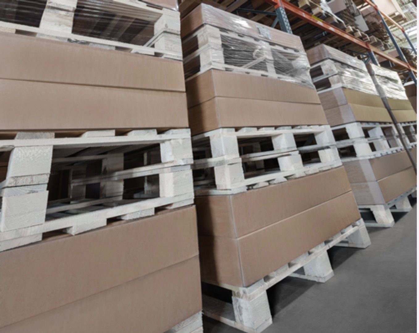 Magazyn: stosy kartonowych pudeł na paletach, owinięte folią stretch, gotowe do transportu. Widok z perspektywy podłogi, regały magazynowe w tle.