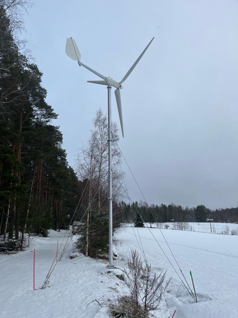 Mała turbina wiatrowa na zasypanym śniegiem terenie, otoczona drzewami i podtrzymywana przez liny. Pochmurne niebo w tle.