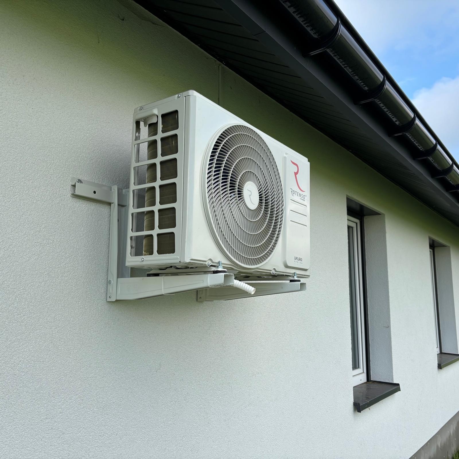 Biała jednostka zewnętrzna klimatyzacji Rotenso Ukura zamontowana na ścianie domu. Widoczne okna i czarny dach. Ujęcie z dołu, lekko z prawej strony.