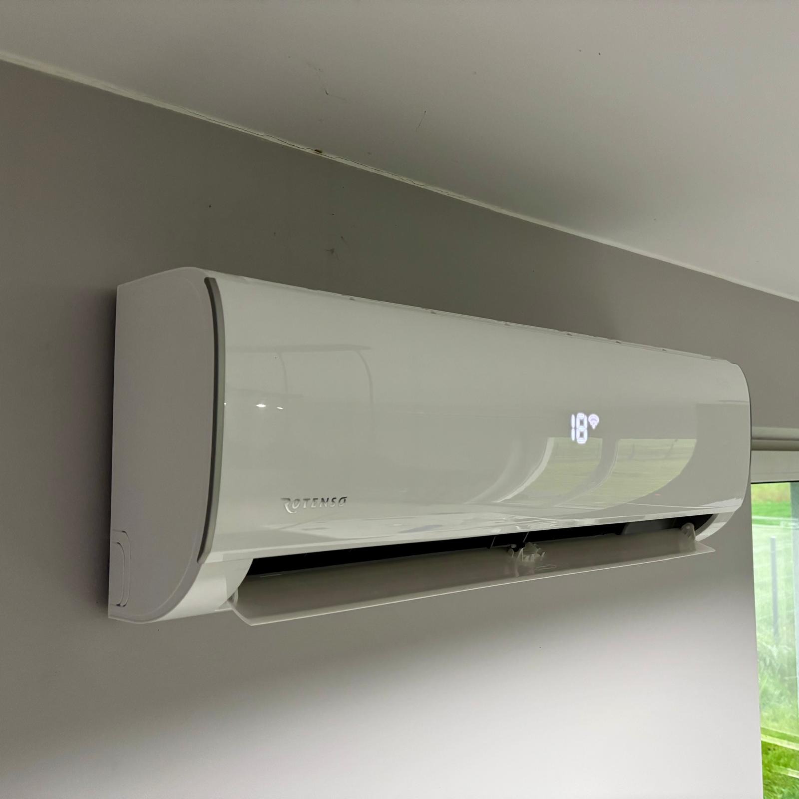 Biały klimatyzator Rotenso zamontowany na szarej ścianie, z wyświetloną temperaturą 18 stopni Celsjusza. Widok z dołu, fragment okna w tle.