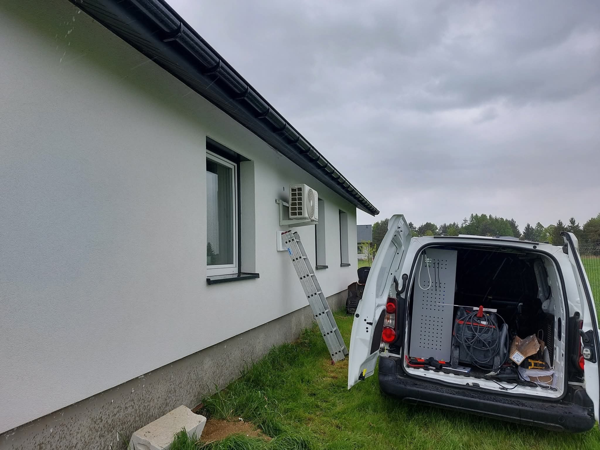 Montaż klimatyzacji na elewacji domu. Drabina oparta o ścianę, otwarty bagażnik samochodu serwisowego z narzędziami w tle, pochmurne niebo.