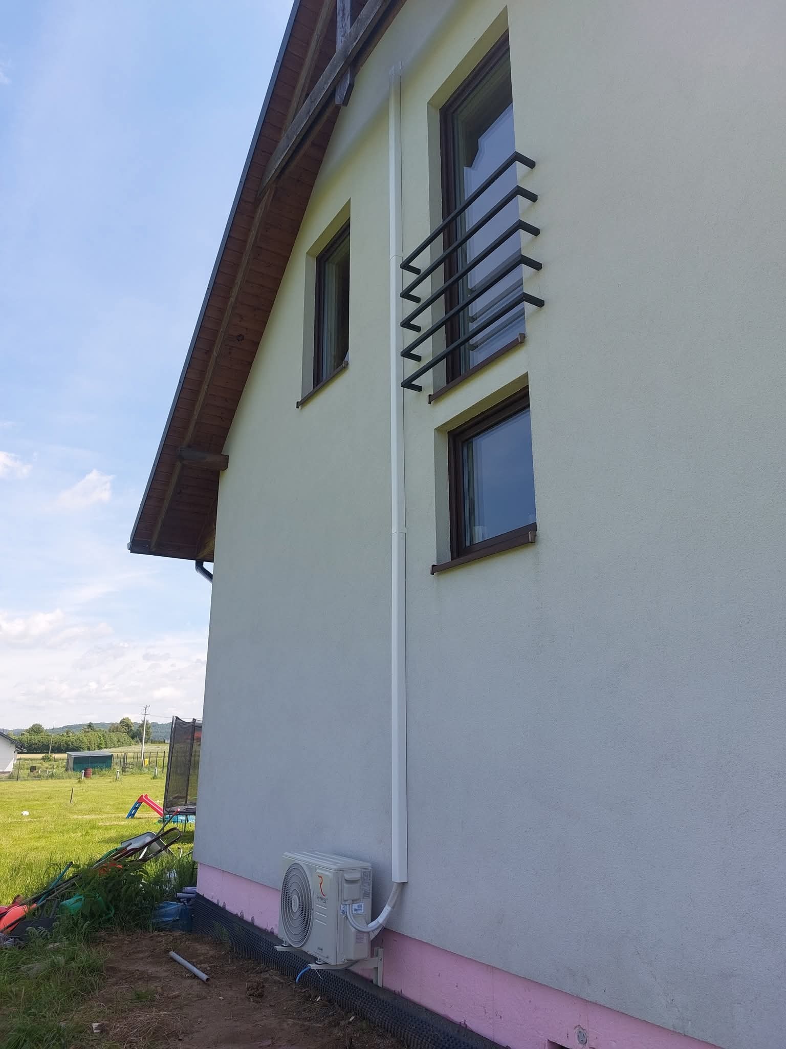 Biała jednostka zewnętrzna klimatyzacji zamontowana na ścianie domu z widoczną instalacją. Tło: trawnik, błękitne niebo.