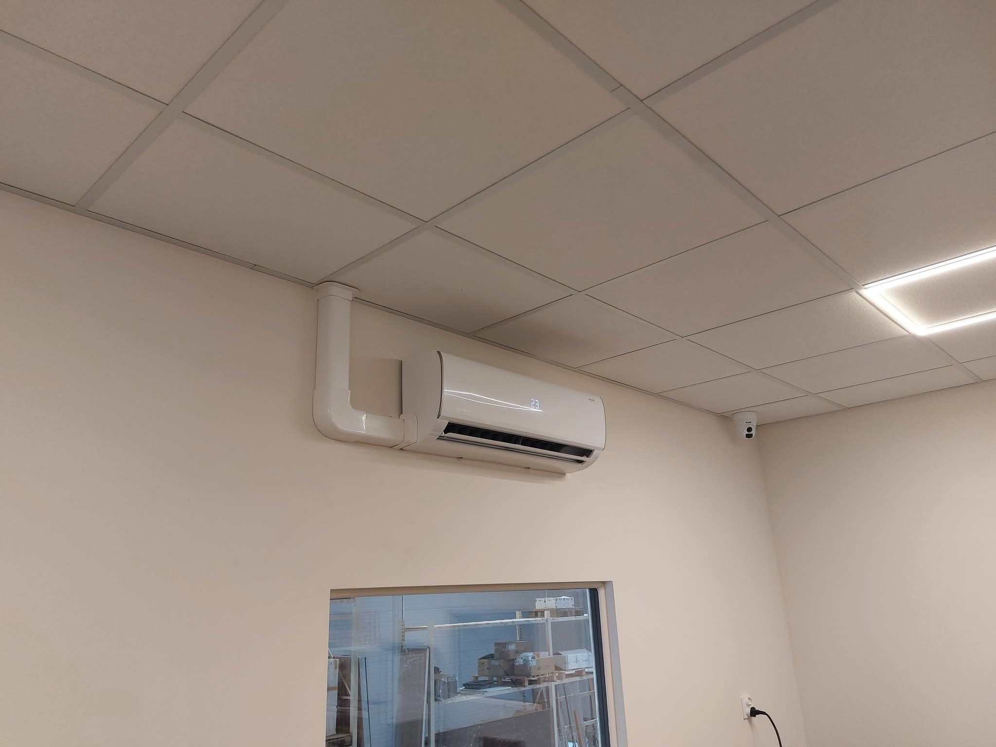 Zamontowany klimatyzator na ścianie w pomieszczeniu z sufitem kasetonowym i oknem. Widoczne rury odprowadzające. Biała obudowa klimatyzatora.
