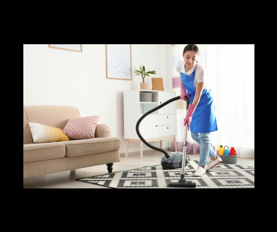 Kobieta w niebieskim fartuchu odkurza wzorzysty dywan w jasnym salonie. W tle sofa, komoda i okno z naturalnym światłem.