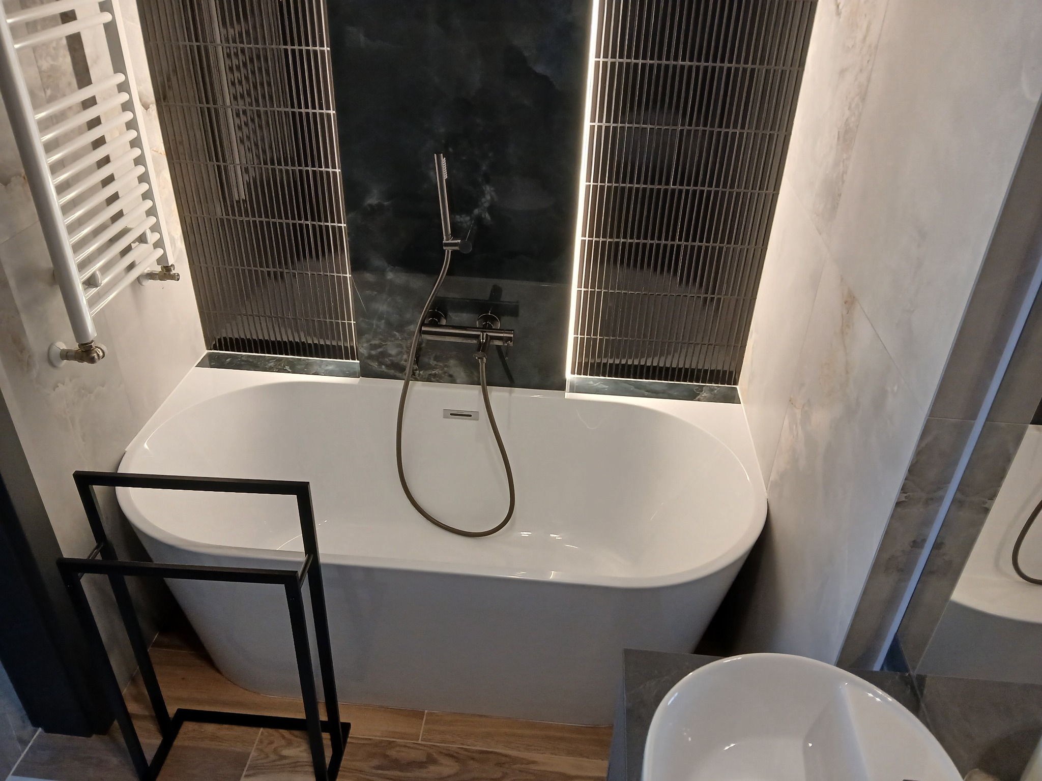 Nowoczesna łazienka z białą wanną, ciemnymi płytkami imitującymi kamień i grzejnikiem drabinkowym. Minimalistyczny design, luksusowe wykończenie.