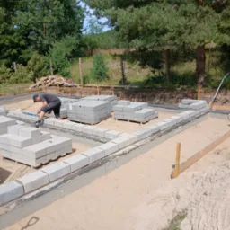 Budowa fundamentów: murarz z poziomicą układa bloczki betonowe na izolacji, w tle drugi pracownik i stos drewna na tle zieleni.