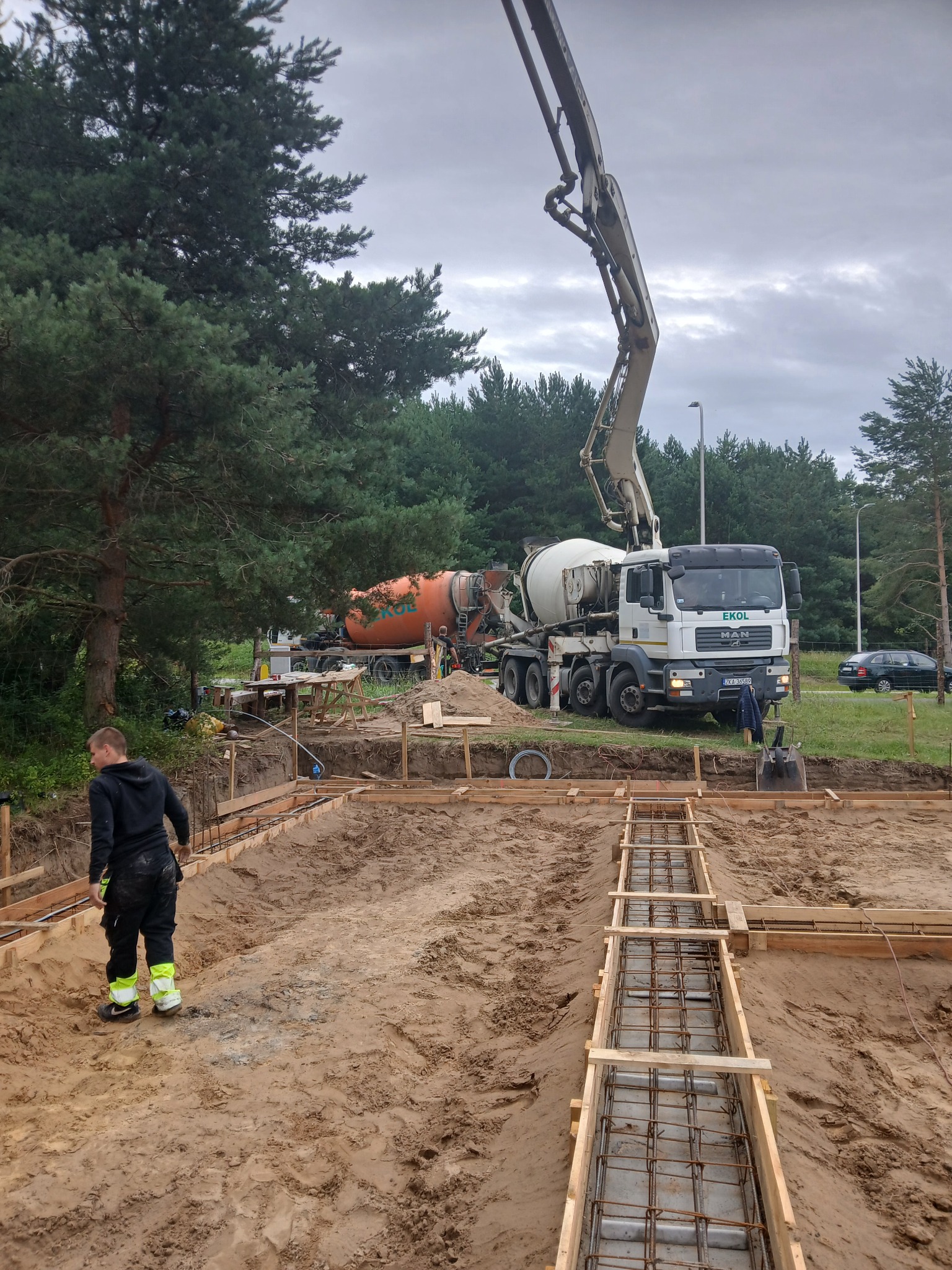 Wykop z szalunkiem i zbrojeniem pod fundament, na tle betonomieszarka z pompą. Widoczny pracownik w odzieży roboczej. Etap budowy domu.
