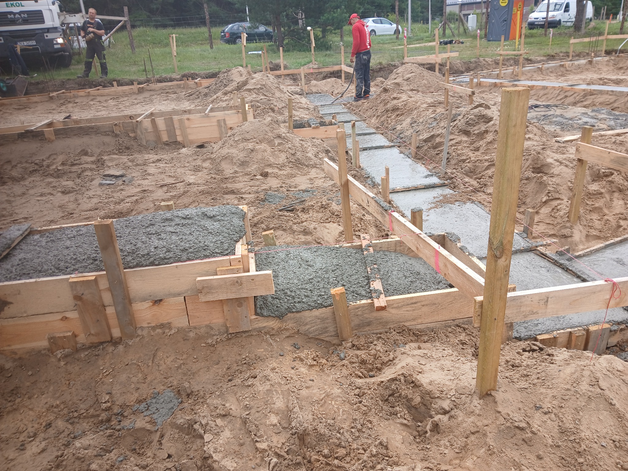 Wypełnione betonem szalunki fundamentowe na placu budowy. Widoczni pracownicy i sprzęt. Ujęcie z poziomu gruntu, podkreślające etap wylewania fundamentów.