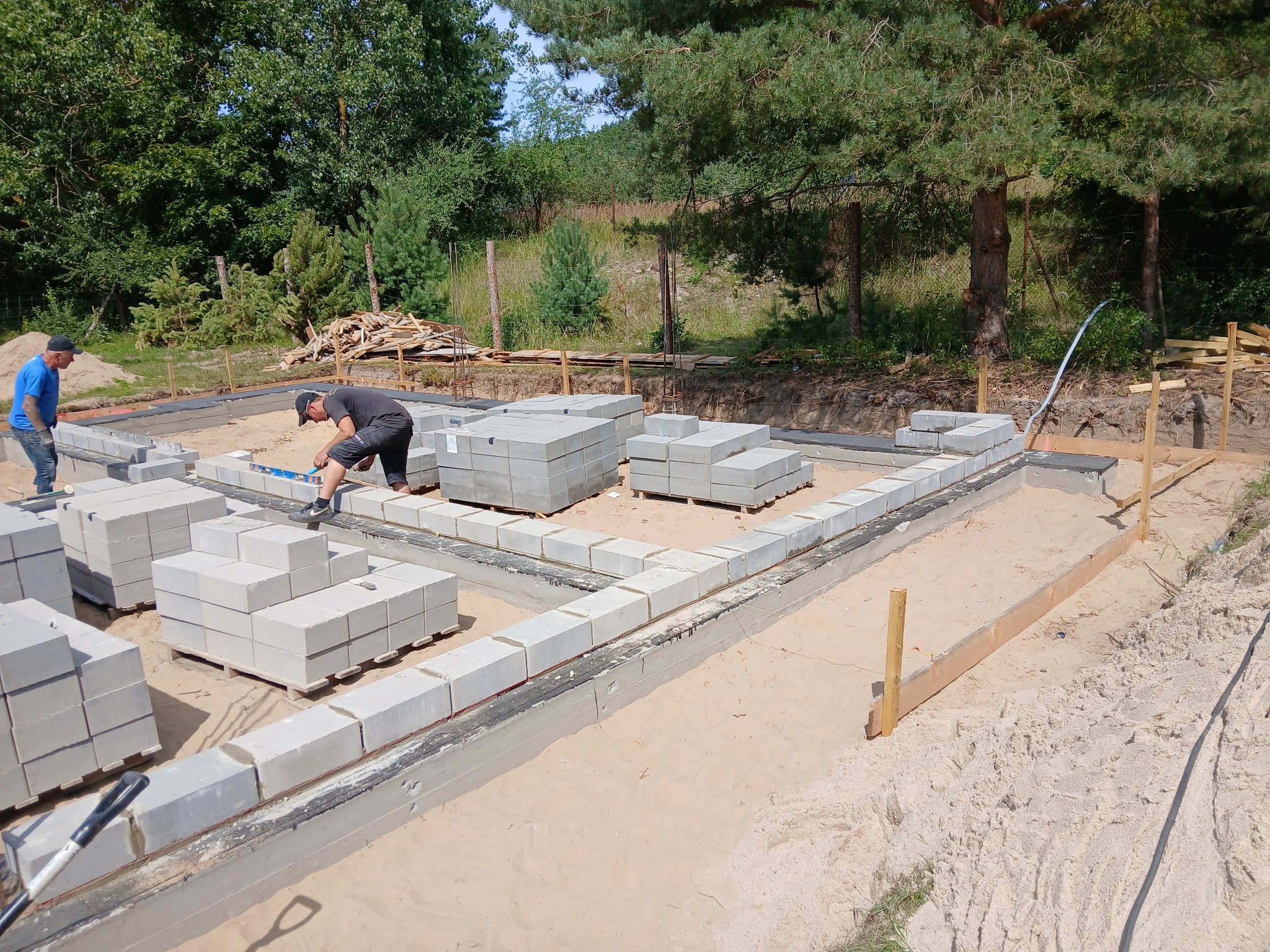 Budowa fundamentów: murarz z poziomicą układa bloczki betonowe na izolacji, w tle drugi pracownik i stos drewna na tle zieleni.