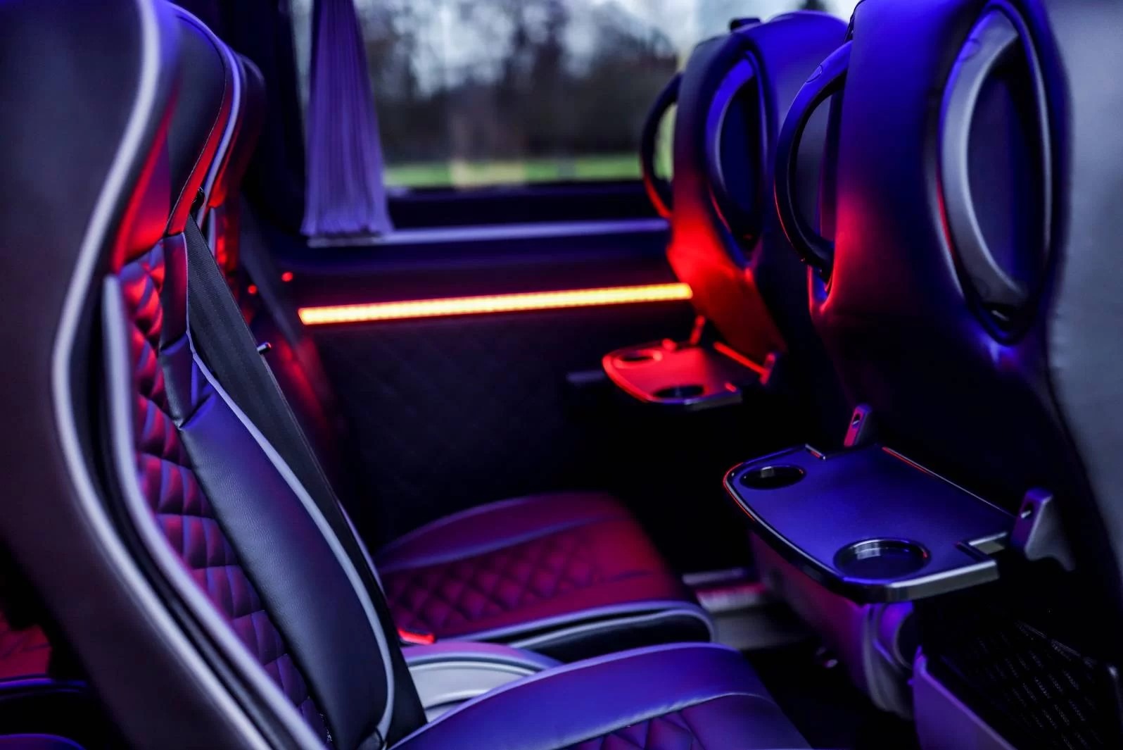 Wnętrze luksusowego busa: czarne skórzane fotele z pikowaniem, białe lamówki, rozkładane stoliki z uchwytami na napoje, oświetlenie LED w kolorach czerwieni i fioletu.