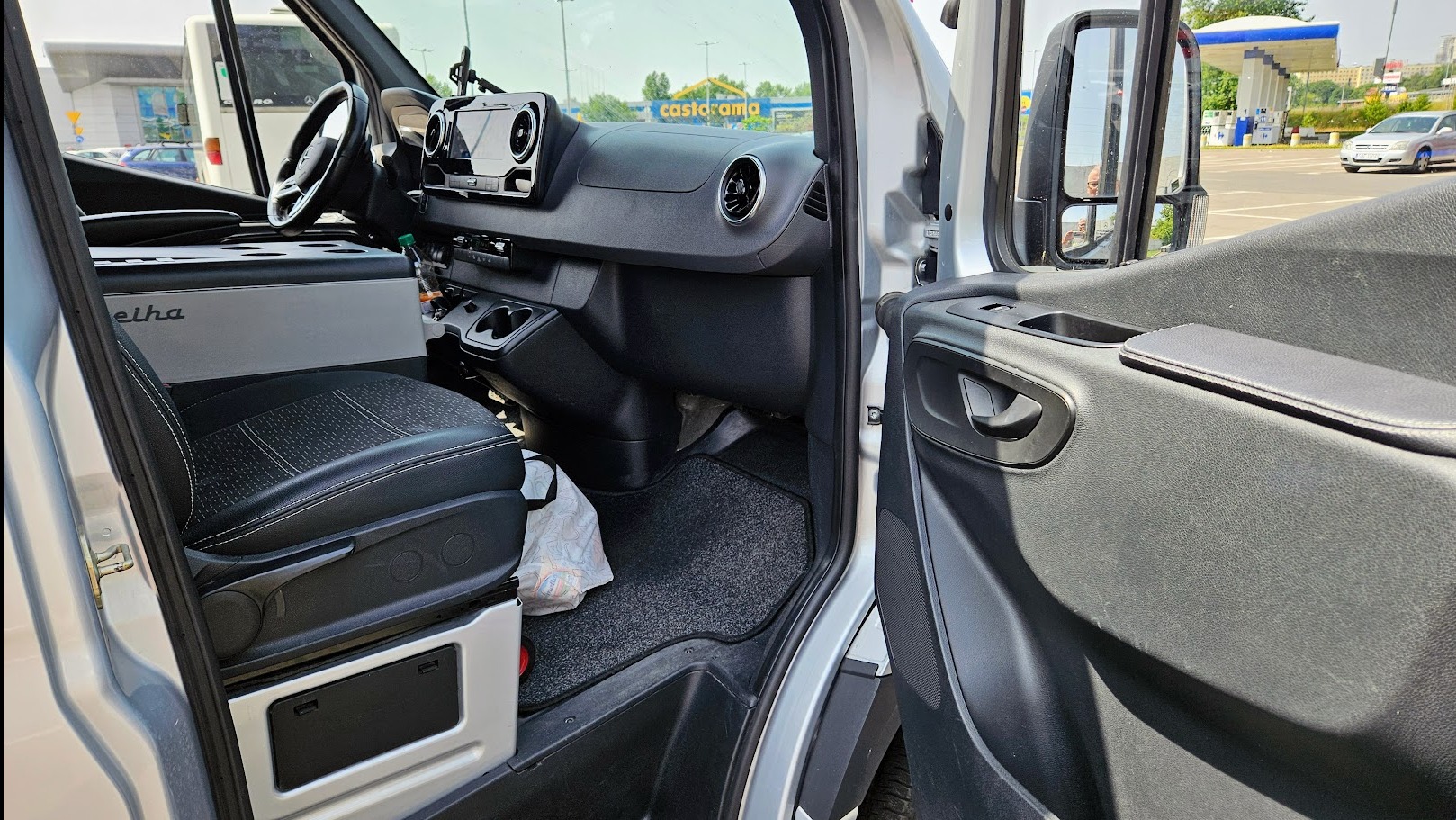 Wnętrze srebrnego busa z otwartymi drzwiami pasażera, widoczny fotel kierowcy, chłodziarka i deska rozdzielcza. W tle parking z Castoramą i stacją benzynową.