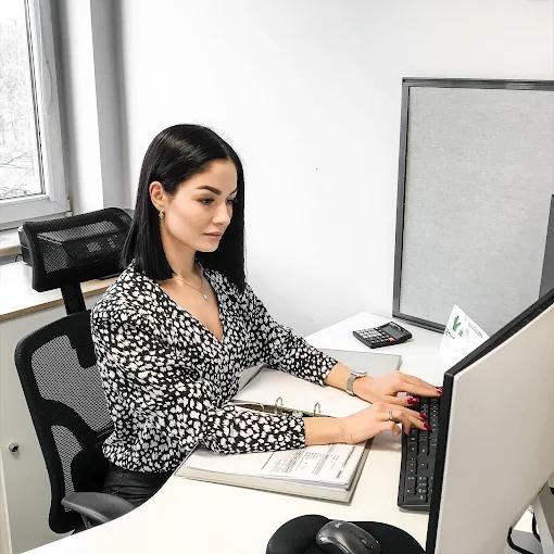 Kobieta w bluzce w cętki, siedząca przy biurku z kalkulatorem i segregatorami, pisze na klawiaturze komputera. Jasne biuro, naturalne światło.