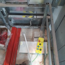 3xNC elektro - Wnętrze szybu windowego w trakcie prac instalacyjnych. Widoczne elementy konstrukcyjne, przewody elektryczne, rury w izolacji i panel sterowania.