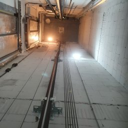 3xNC elektro - Wnętrze szybu windowego w trakcie budowy, widoczne szyny prowadzące, kable i oświetlenie robocze. Ściany z białych bloków, sufit z instalacjami.