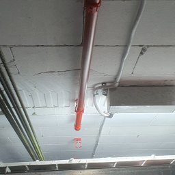 3xNC elektro - Instalacja rur i kabli na suficie z bloków betonowych, widok z dołu. Czerwona rura pionowo, szare kable poziomo. Metalowa konstrukcja w dolnej części kadru.