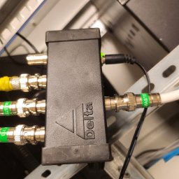 3xNC elektro - Czarny rozdzielacz Delta z podłączonymi kablami BNC i zasilaniem w szafie rack, widoczne oznaczenia 'MASTER' na kablach. Element instalacji alarmowej lub monitoringu.