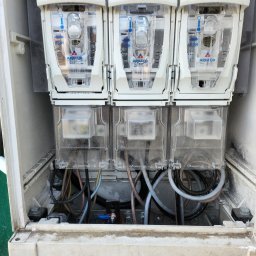 3xNC elektro - Otwarta skrzynka z trzema licznikami energii elektrycznej marki Apator, widoczne przewody i elementy montażowe wewnątrz. Obudowa lekko zabrudzona.