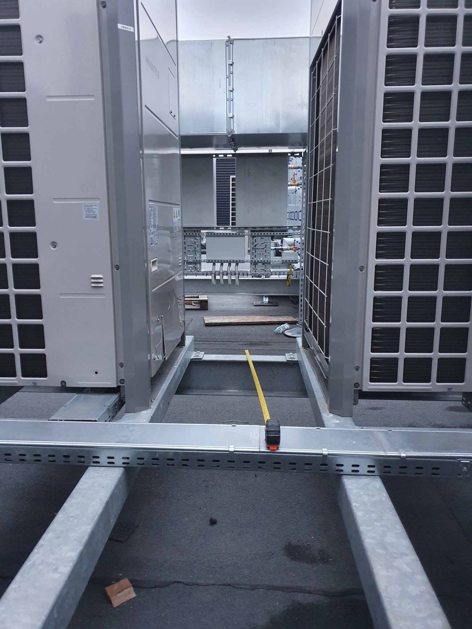 Widok na moduły przemysłowej instalacji klimatyzacji, z widoczną taśmą mierniczą, sugerującą pomiary. Szara kolorystyka. Metalowe konstrukcje.