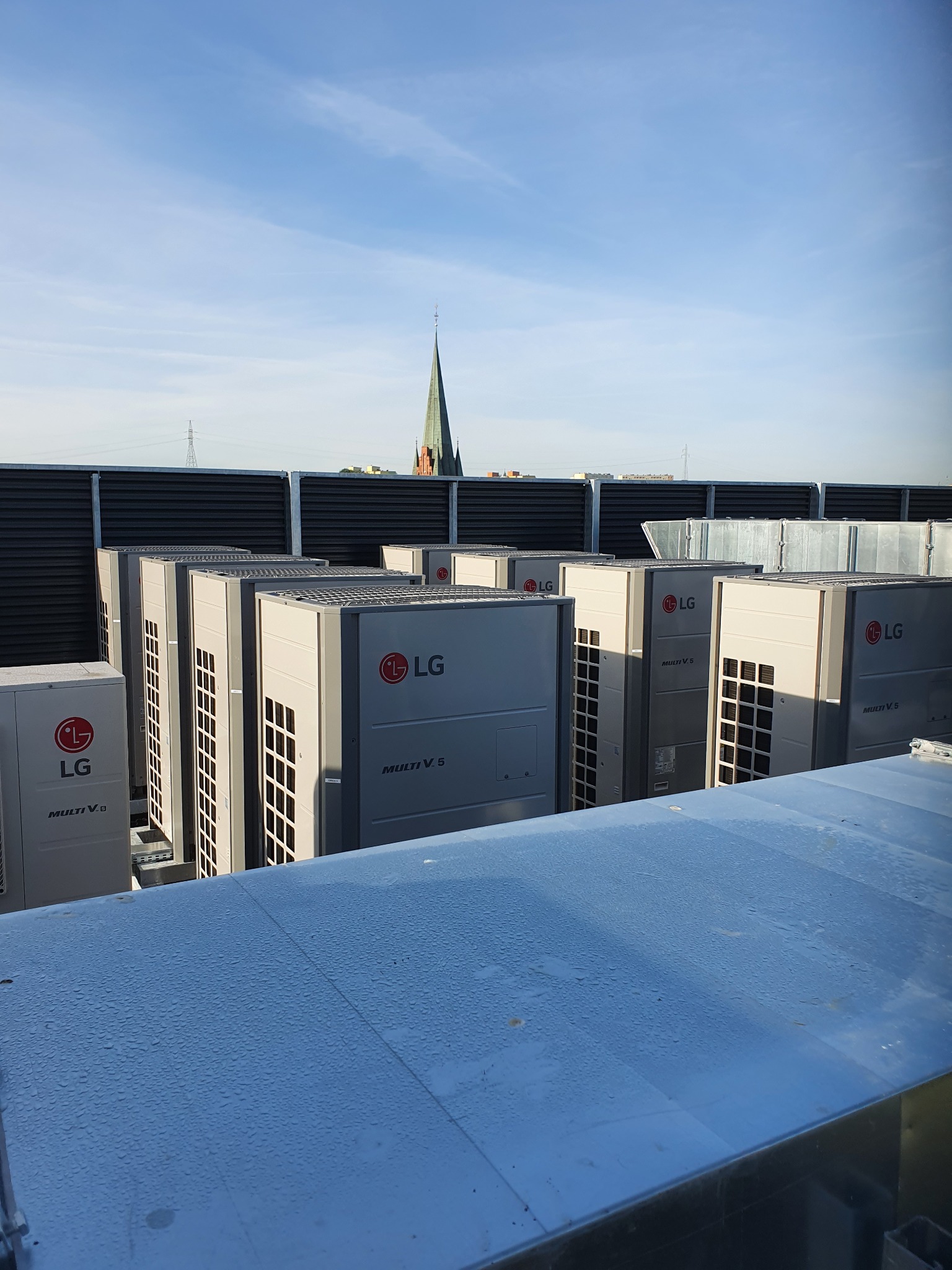 Rząd jednostek zewnętrznych klimatyzacji LG Multi V 5 na dachu budynku, z widokiem na iglicę kościoła i błękitne niebo w tle. Instalacja komercyjna.