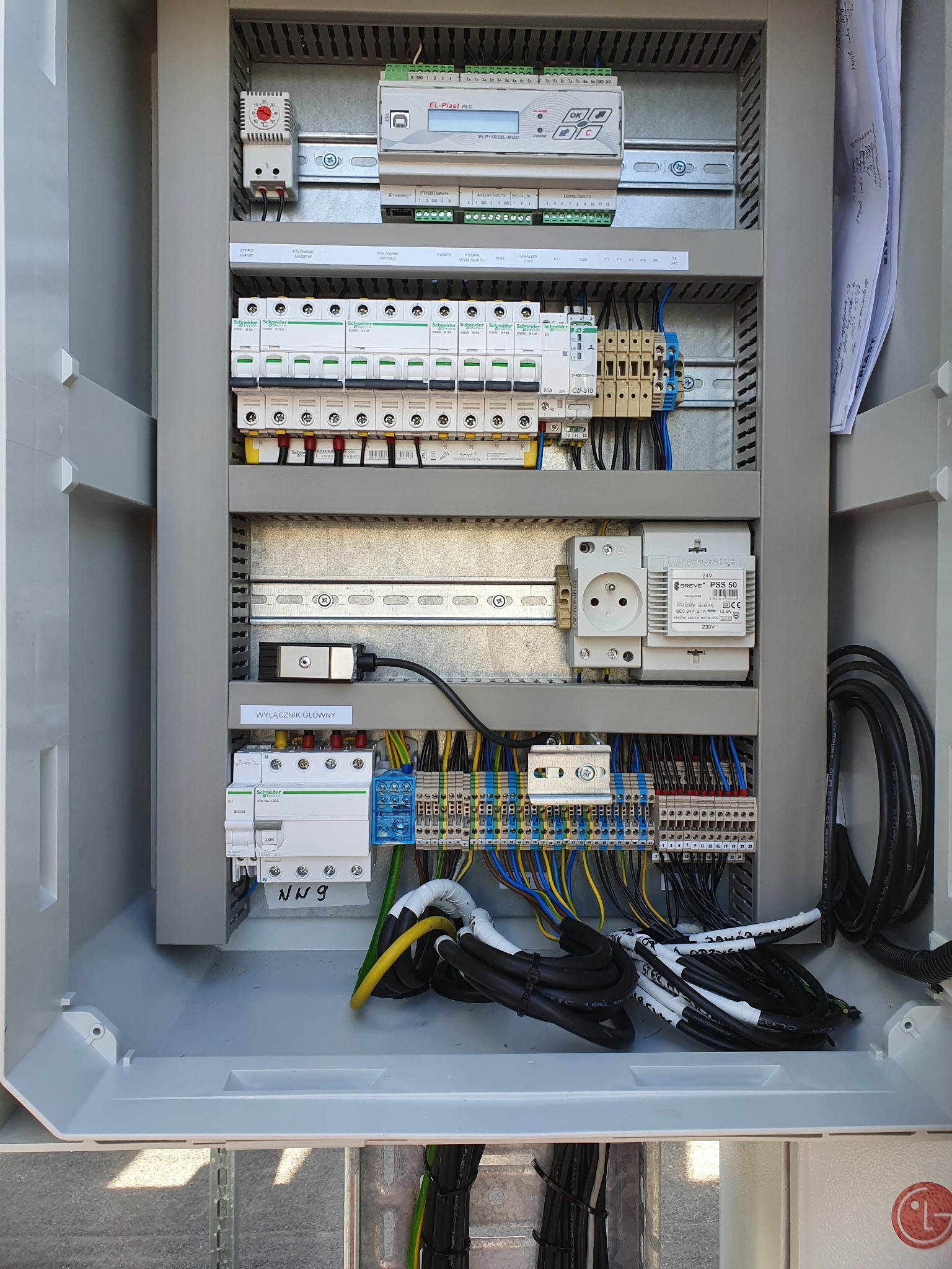 Otwarta, szara szafa sterownicza z widocznymi bezpiecznikami, przewodami i elementami sterującymi. Widoczne oznaczenia i etykiety na przewodach.