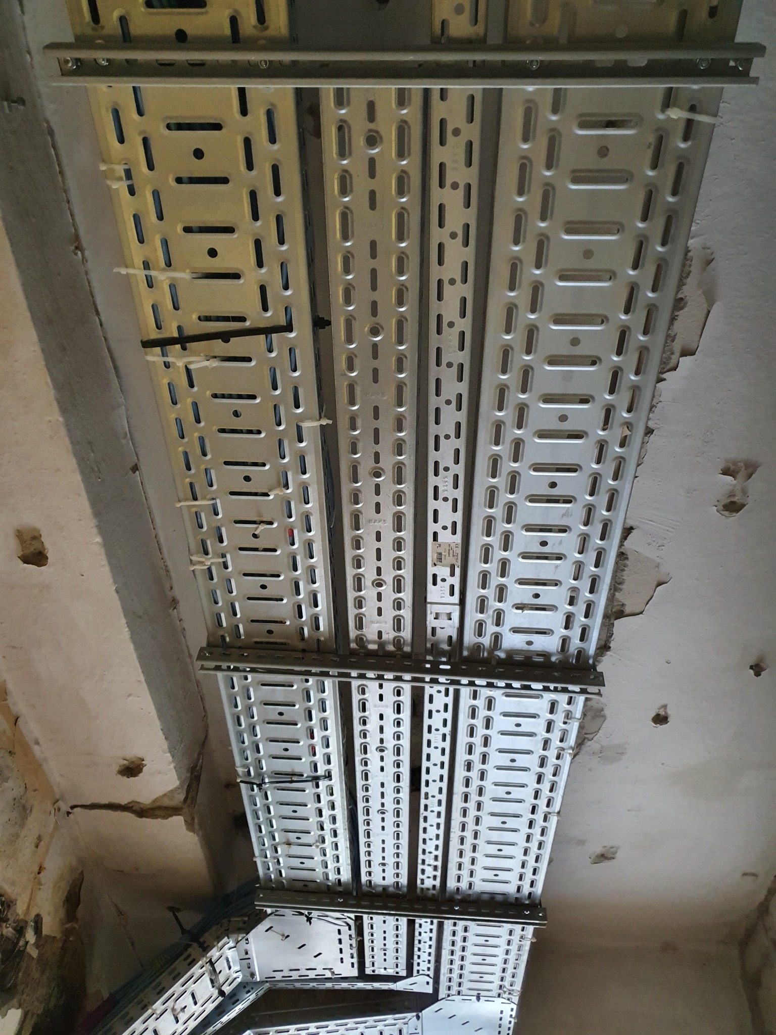 Metalowe korytka kablowe zamontowane do sufitu w surowym stanie, widoczne przewody zabezpieczone opaskami. Instalacja elektryczna w trakcie realizacji.