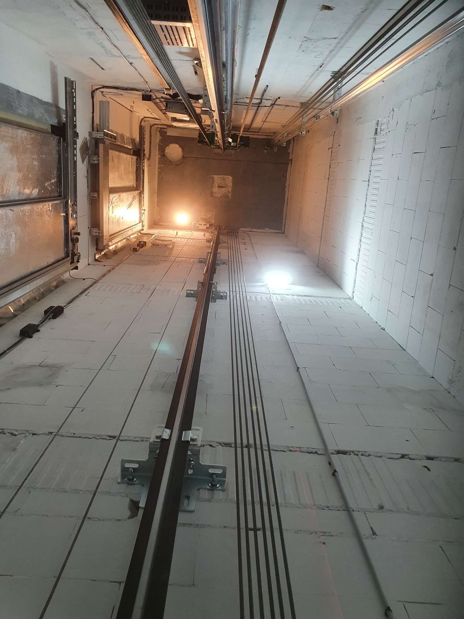 Wnętrze szybu windowego w trakcie budowy, widoczne szyny prowadzące, kable i oświetlenie robocze. Ściany z białych bloków, sufit z instalacjami.
